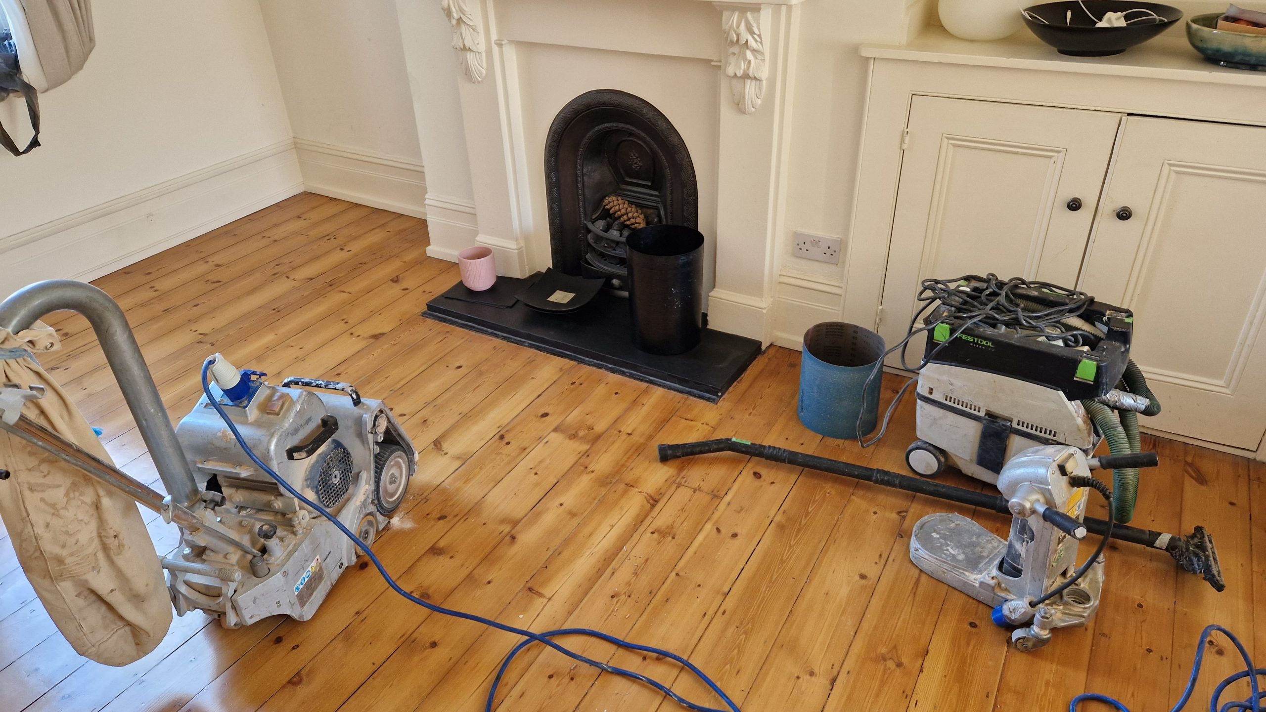  Wood Floor Sanding tottenham scene showing freshly sanded solid pine floorboards in a Tottenham living room, with drum and edge sanders plus a dust-extraction unit beside a cast-iron fireplace.